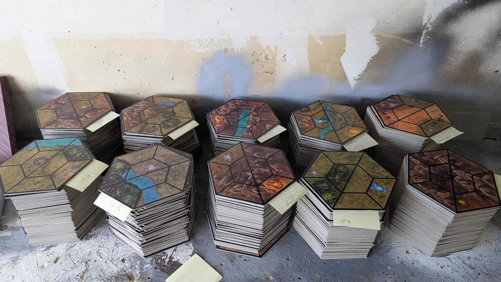 UV-printed game boards and modular hex tiles by Kulworks: navy and gold board panels printing on the flatbed and coming off finished, hex terrain tiles cut on the clicker press and freed from the die, a full run stacked and bundled on the bench, and tile artwork prepped for print